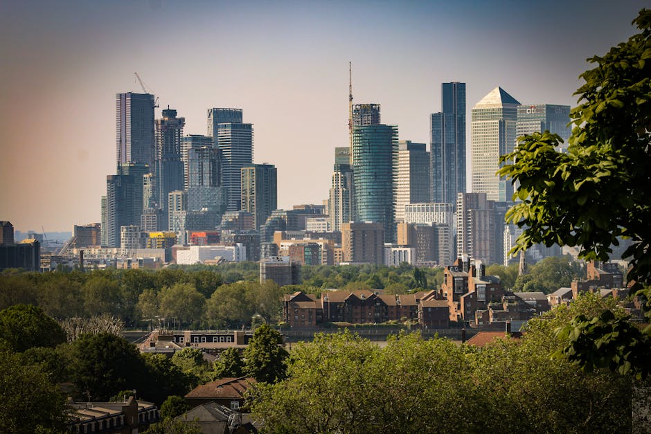 A cityscape featuring a dense cluster of modern skyscrapers with glass facades and varying heights, set against a light sky with subtle cloud cover. In the foreground, there is a lush greenery scene i