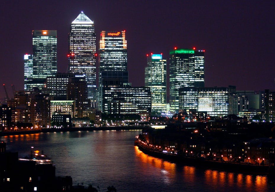 A cityscape featuring a dense cluster of modern skyscrapers with glass facades and varying heights, set against a light sky with subtle cloud cover. In the foreground, there is a lush greenery scene i