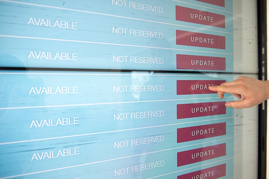 A person's hand is seen pointing at a red 'Update' button on a digital screen displaying a booking schedule with multiple rows. Each row shows the status 'Available' and 'Not Reserved' next to the but
