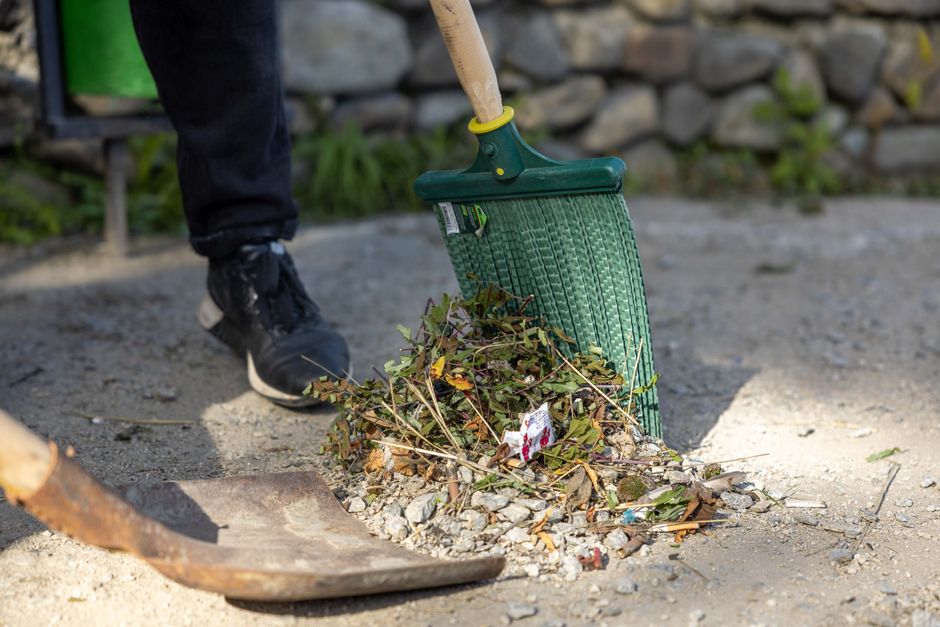A person wearing black footwear and dark pants is using a green push broom with a yellow connection to sweep dry leaves, twigs, and small debris into a pile on a gravel surface. The scene appears to b